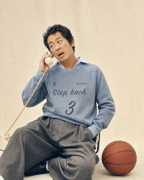 A man sitting on a chair holding a vintage cream-colored telephone receiver to his ear, wearing a light blue sweatshirt with 'Step back 3' printed on the front and loose gray sweatpants. A basketball sits on the floor next to him against a warm beige background.